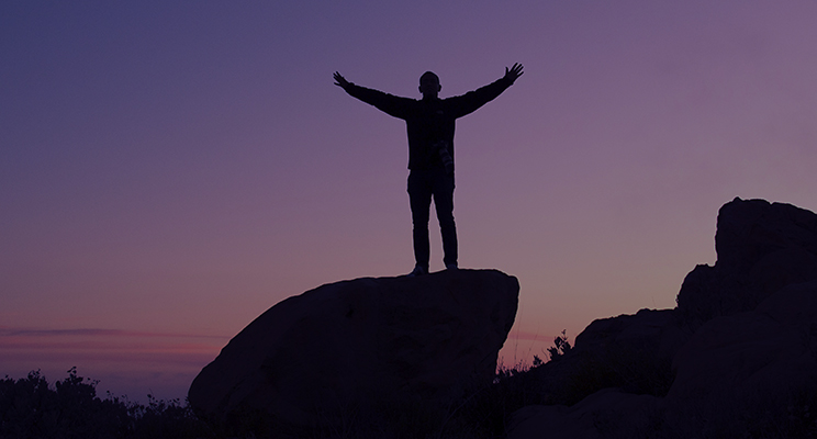 Silhouette of person with out stretched arms standing on a rock at dusk